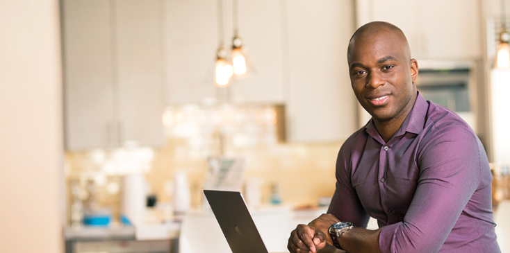 A man in a purple dress shirt works on his laptop with a kitchen behind him