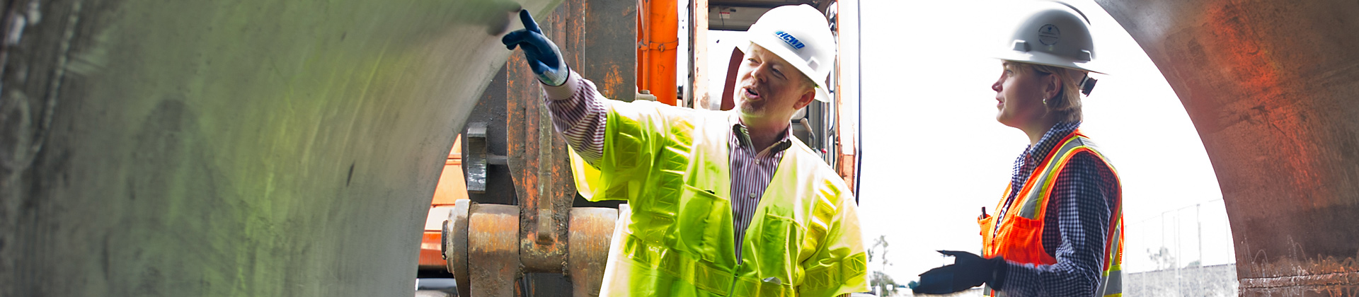 A man and a woman in hard hats and safety vests look at a giant pipe