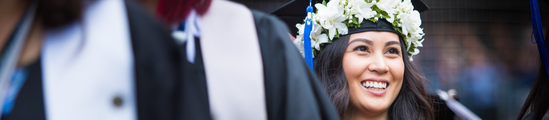 woman in cap and gown smiling off to her left wearing flowers around graduation cap after earning her bachelor's degree