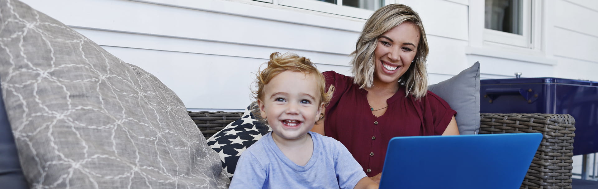 A mother sits on a couch with a laptop and smiles at her toddler-aged son, who is looking forward.