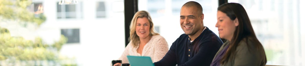 three individuals a woman, man and woman sit in office at table looking down at laptop smiling