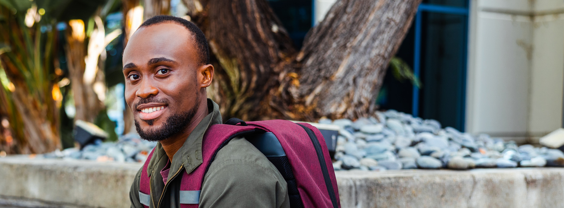 Man earning associate's degree is wearing a burgundy backpack and smiles at camera.