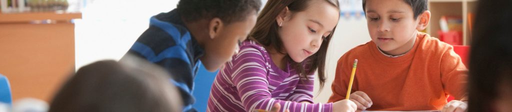 3 elementary school aged children, a girl writes with a pencil with boys on either side looking down at her paper