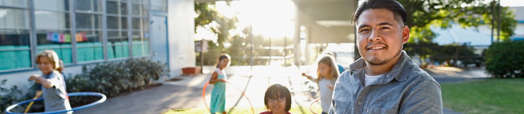 man outside smiling with young elementary school kids playing in background