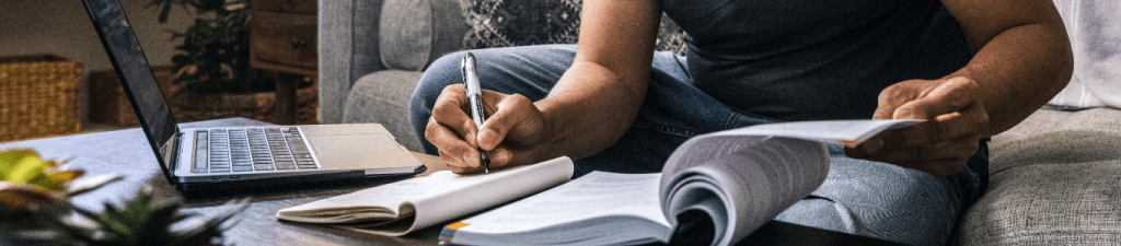 man sitting on couch taking notes as he flips through textbook