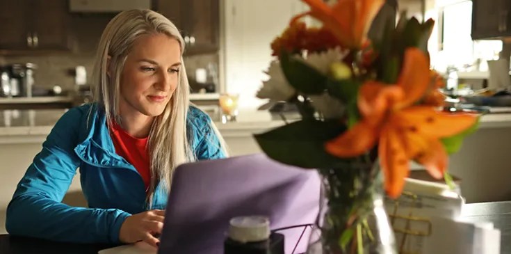 woman sitting at kitchen table with purple laptop and orange flowers